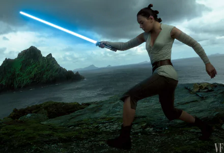 Rey from Star Wars: The Last Jedi wields a blue lightsaber on a rocky coastline under a cloudy sky. This HD desktop wallpaper features Daisy Ridley's character in an iconic pose.