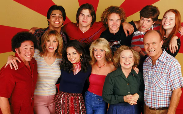 HD PC desktop wallpaper showing a smiling ensemble from a 1970s‑set TV sitcom posed against a red-and-yellow sunburst background.