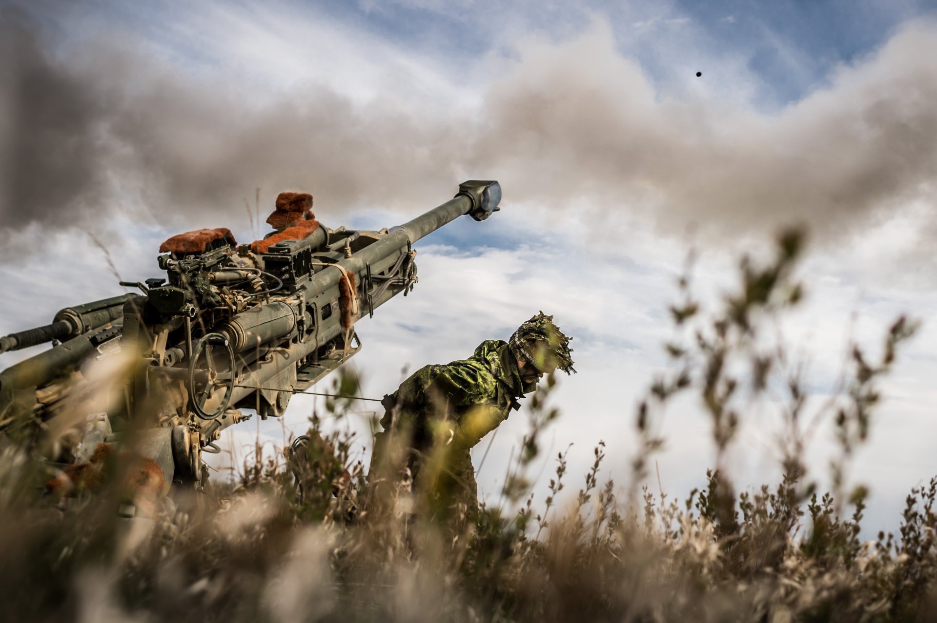 4K Ultra HD PC desktop wallpaper showing military artillery: low-angle view of a heavy weapon and camouflaged soldier firing through smoke and tall grass.