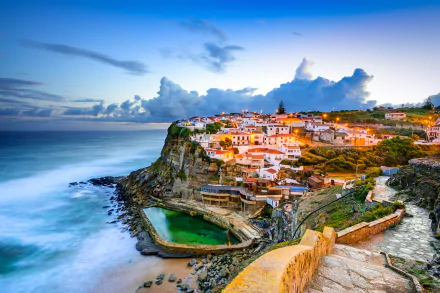 A stunning view of Azenhas do Mar in Sintra, Portugal, with a coastal town perched on cliffs overlooking the ocean, vibrant sky, cloud formations, and charming houses illuminated as evening falls. HD desktop wallpaper.