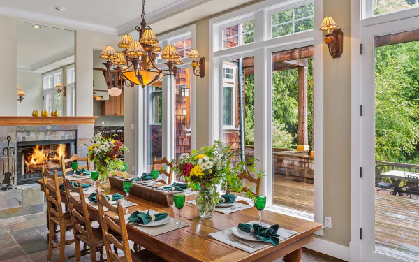 HD PC desktop wallpaper: bright man-made dining room interior with wooden table and chairs, lit fireplace, chandelier, large windows and doors opening to a green patio; decorative table settings.