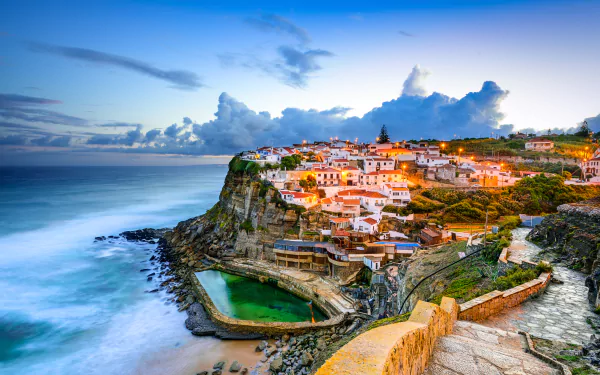 A stunning view of Azenhas do Mar in Sintra, Portugal, with a coastal town perched on cliffs overlooking the ocean, vibrant sky, cloud formations, and charming houses illuminated as evening falls. HD desktop wallpaper.