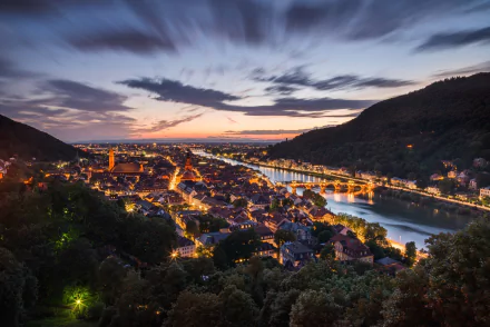 Nighttime HD landscape of Heidelberg, Germany, featuring a river winding through the illuminated town nestled between hills, showcasing a vibrant man-made scene.