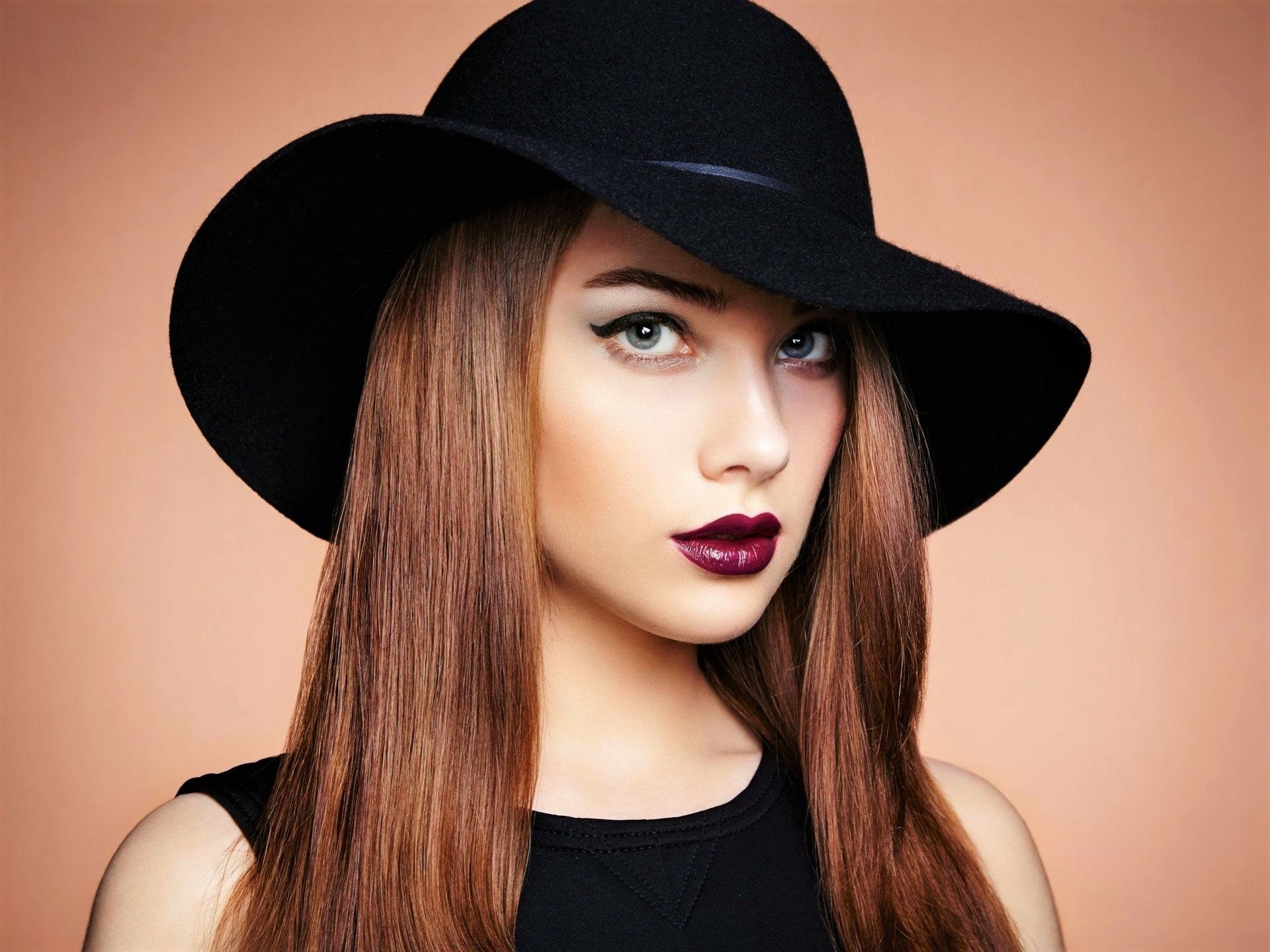 HD desktop wallpaper of a woman model with blue eyes, wearing dark lipstick and a black fedora hat, against a soft peach background.