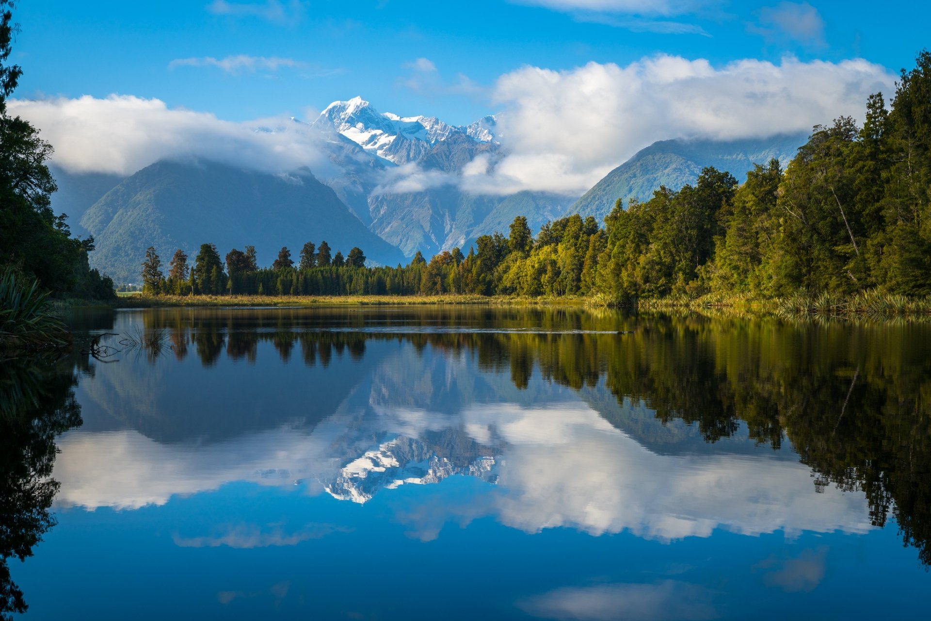 HD desktop wallpaper showing Aoraki/Mount Cook reflected in Lake Matheson with clouds and lush greenery in New Zealand's natural landscape.