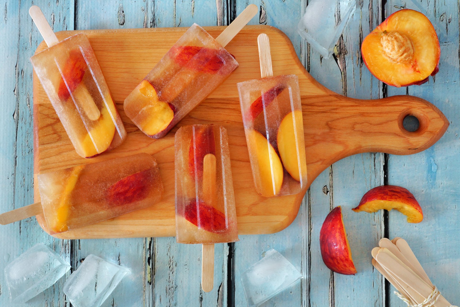 Colorful fruit ice cream popsicles with visible peach slices and ice cubes rest on a wooden cutting board, set against a rustic blue background in vibrant 4K Ultra HD detail.