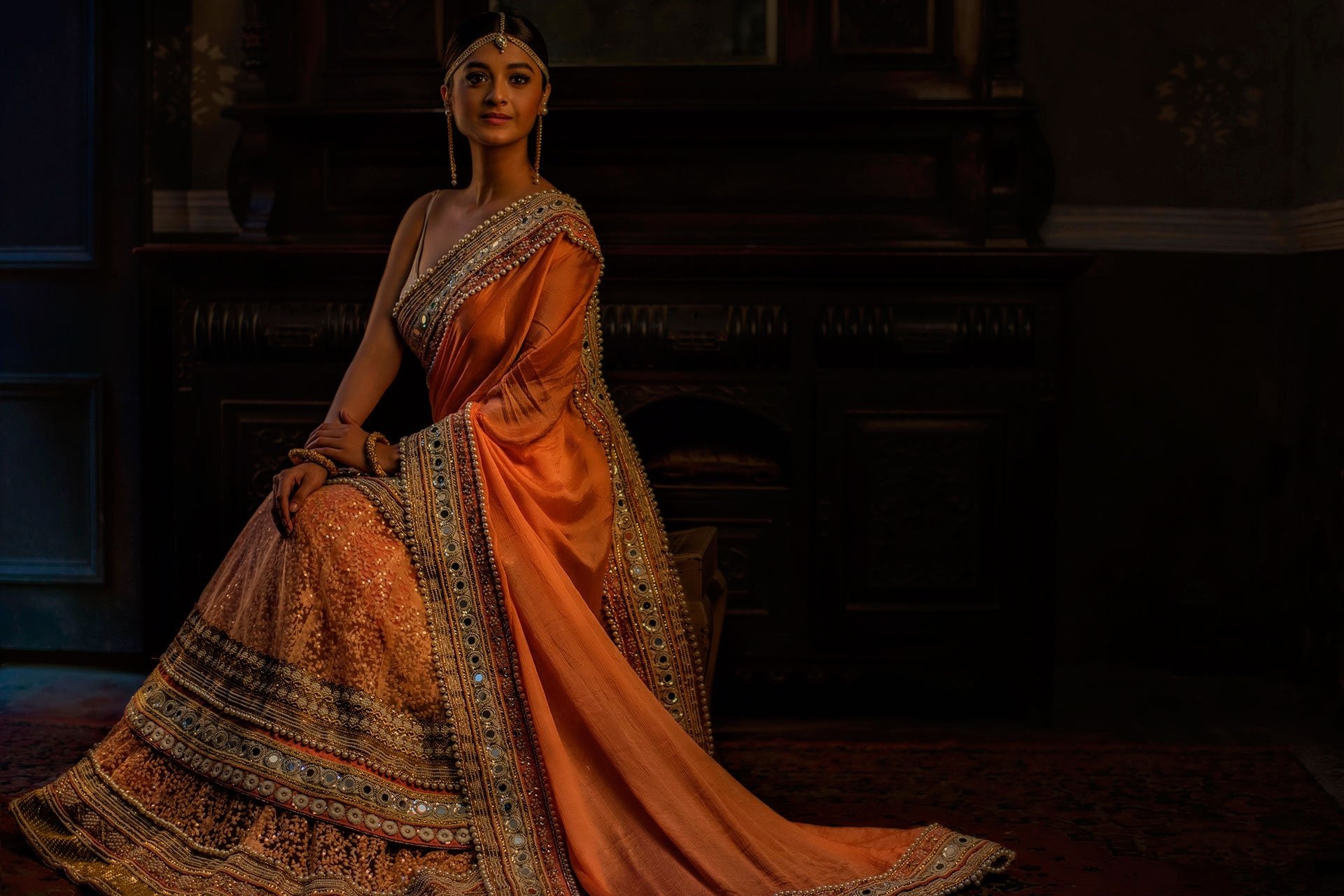 An artistic HD wallpaper featuring a brown-eyed Indian woman adorned with jewelry and wearing a beautifully detailed saree. She sits gracefully, exuding elegance and tradition in an oriental setting.