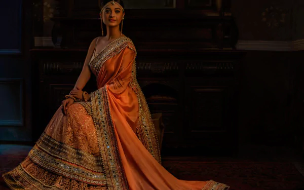 An artistic HD wallpaper featuring a brown-eyed Indian woman adorned with jewelry and wearing a beautifully detailed saree. She sits gracefully, exuding elegance and tradition in an oriental setting.