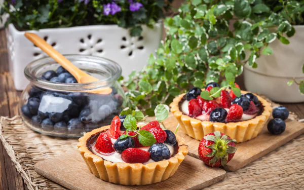 Close-up of blueberry and strawberry tarts with fresh berries and mint leaves on a wooden surface, surrounded by green plants, captured in 4K Ultra HD.