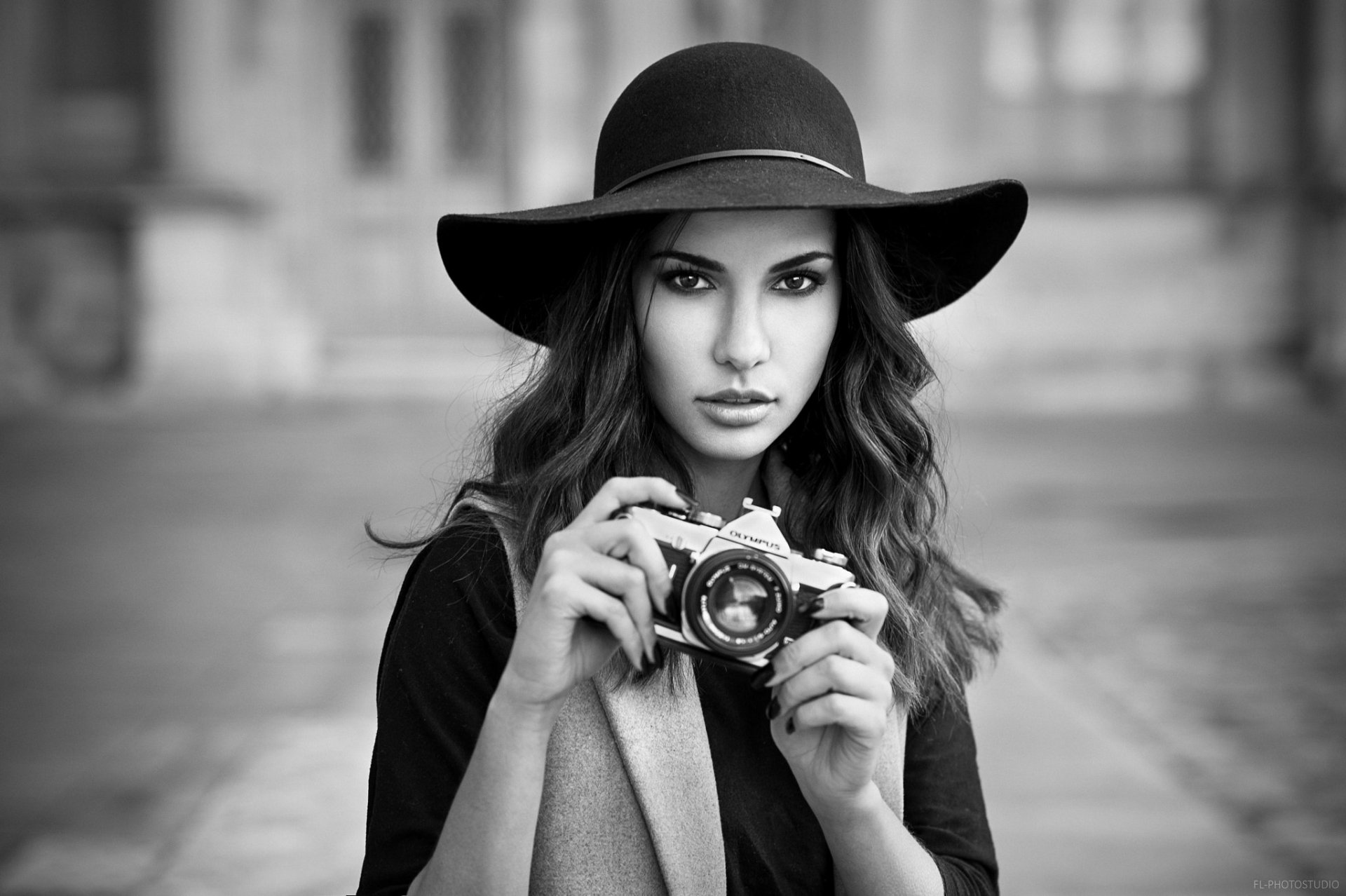 Black & white HD desktop wallpaper: female model in a wide-brim hat holding an Olympus camera, intense stare, shallow depth of field and blurred urban background.