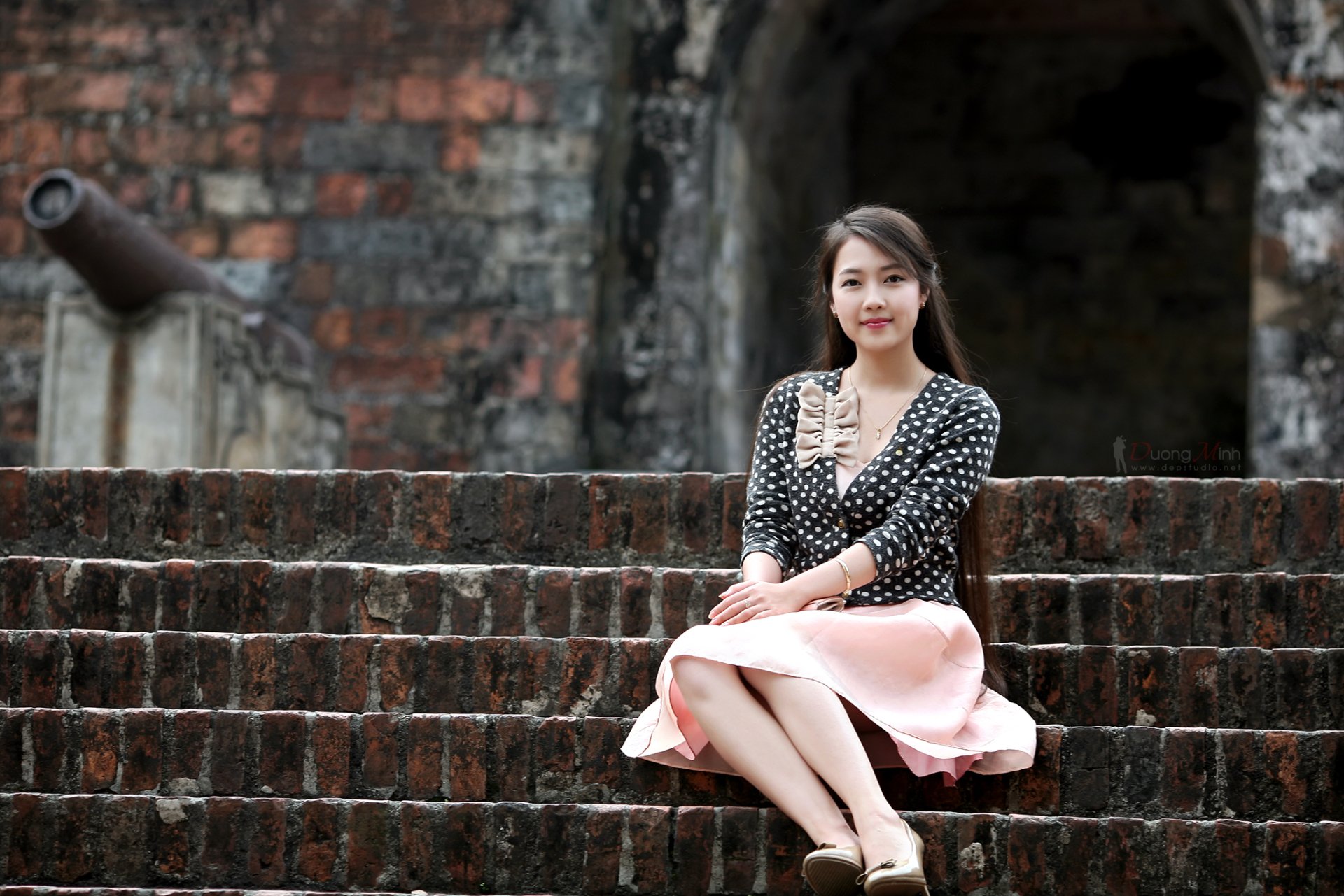 A smiling brunette Asian woman sits on stone steps in front of an ancient wall, creating a serene and picturesque HD desktop wallpaper.