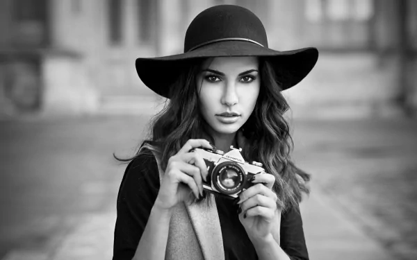 Black & white HD desktop wallpaper: female model in a wide-brim hat holding an Olympus camera, intense stare, shallow depth of field and blurred urban background.