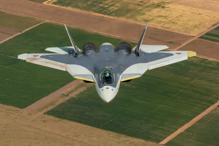 A Sukhoi Su-57 military jet fighter warplane flying over patchwork farmland, captured in high-definition detail as a PC desktop wallpaper.