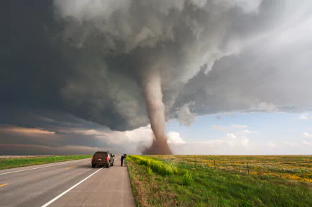 HD desktop wallpaper of a tornado touching down in a vast green field under dark storm clouds, with a road and parked vehicles in the foreground.