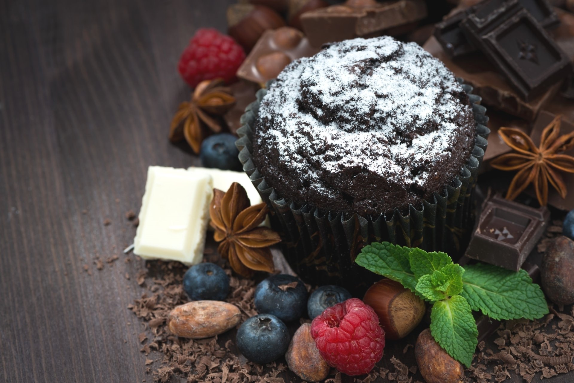 HD still life wallpaper featuring a chocolate muffin surrounded by star anise, chocolate pieces, raspberries, blueberries, and fresh mint on a wooden surface.