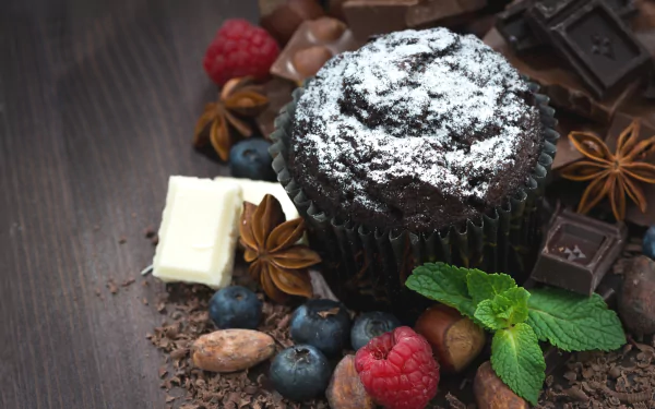 HD still life wallpaper featuring a chocolate muffin surrounded by star anise, chocolate pieces, raspberries, blueberries, and fresh mint on a wooden surface.