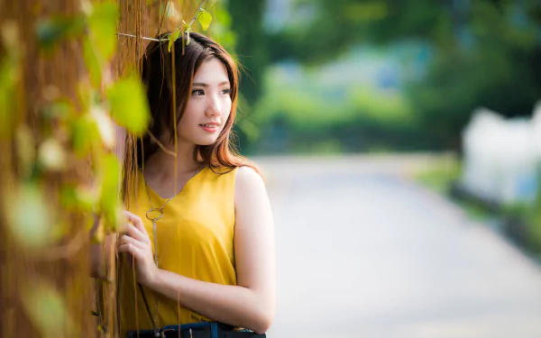 A brunette Asian woman with brown eyes poses thoughtfully by a wall, captured in sharp focus with a soft, blurred background in 8K Ultra HD quality.