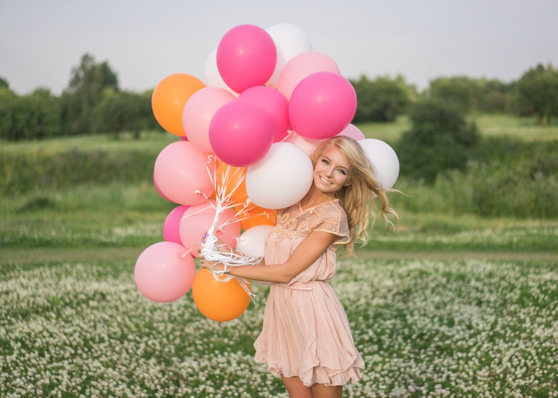 A smiling blonde woman with blue eyes in a pink dress holds colorful balloons in a field, captured with a soft depth of field in this HD desktop wallpaper.