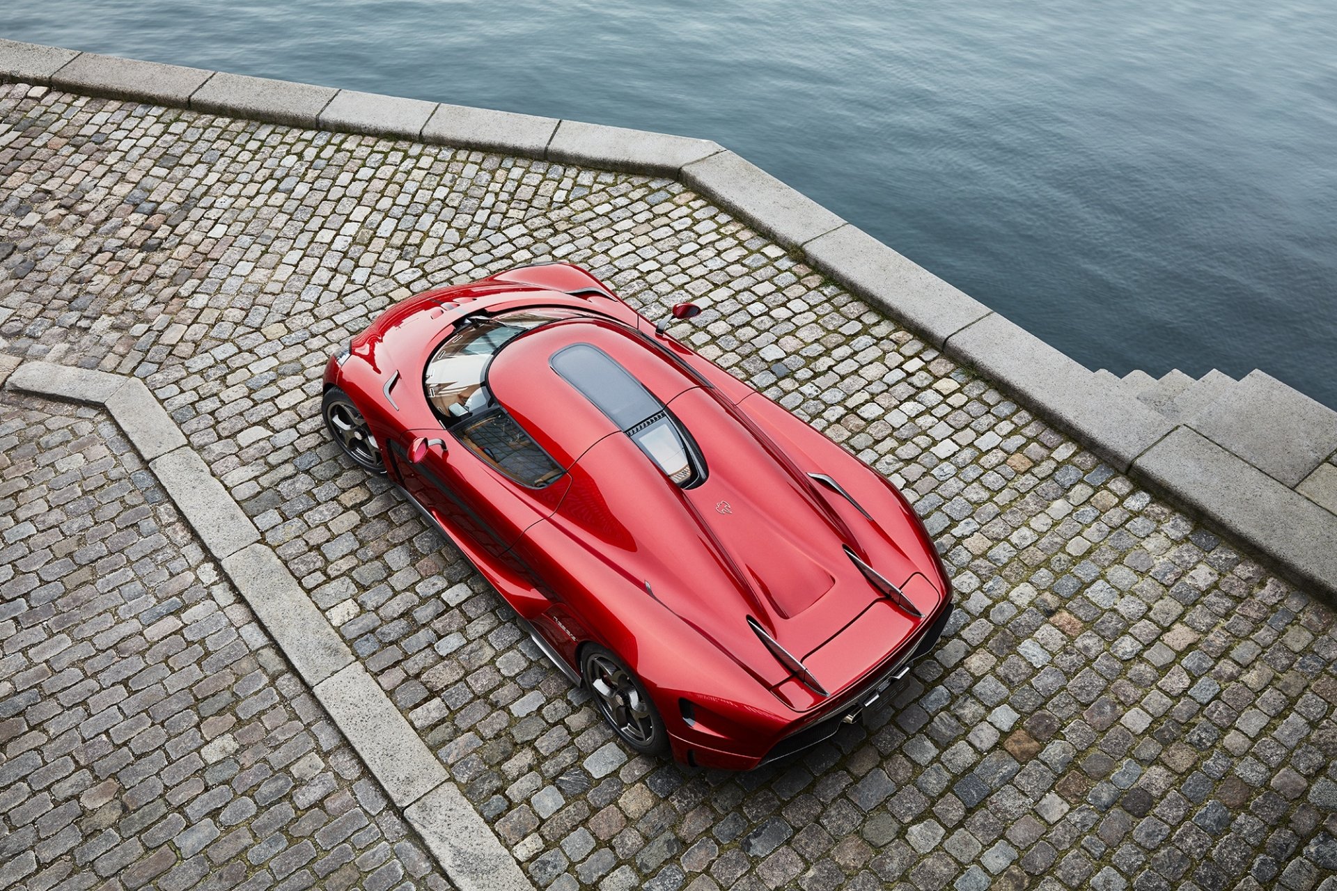 HD PC desktop wallpaper and background: red Koenigsegg Regera supercar car, a sleek Koenigsegg vehicle parked on cobblestone quay beside the water, aerial perspective.