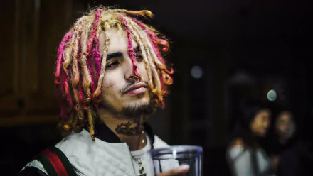 HD desktop wallpaper of hip hop artist Lil Pump with colorful dreadlocks, holding a drink, set against a dimly lit background with blurred figures. Tagged: Hip Hop, music.