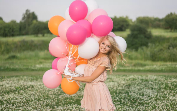 A smiling blonde woman with blue eyes in a pink dress holds colorful balloons in a field, captured with a soft depth of field in this HD desktop wallpaper.
