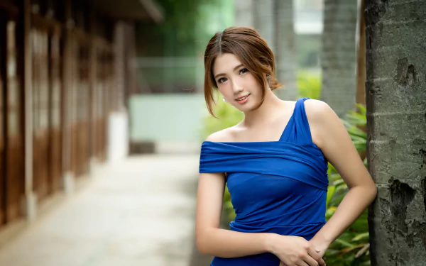 Asian woman in a blue dress smiling, stands leaning on a tree. The background is blurred, showcasing a depth of field effect. The wallpaper is in HD, capturing her brunette hair and model-like appearance.
