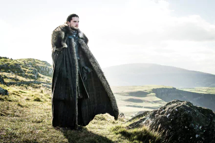 Kit Harington as Jon Snow stands on a grassy hillside with a fur cloak, set against a misty, mountainous landscape in this 4K Ultra HD Game of Thrones wallpaper.