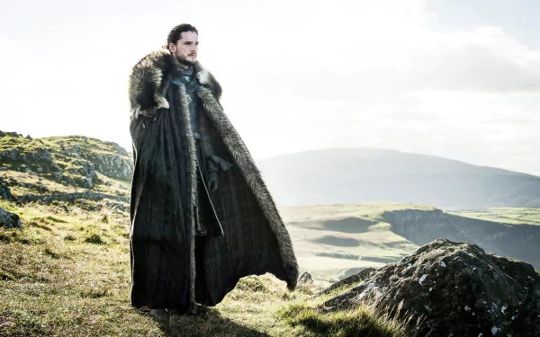 Kit Harington as Jon Snow stands on a grassy hillside with a fur cloak, set against a misty, mountainous landscape in this 4K Ultra HD Game of Thrones wallpaper.