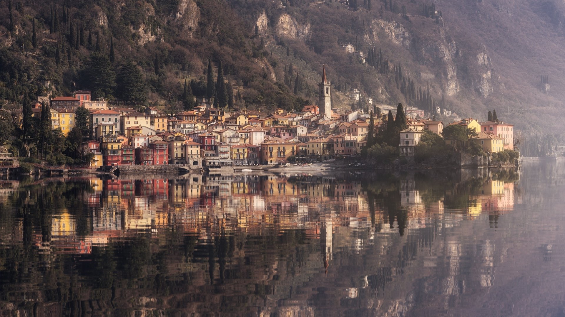 HD desktop wallpaper of Varenna, an Italian village by a lake, showcasing colorful houses and their reflection on the calm water beneath rocky cliffs.