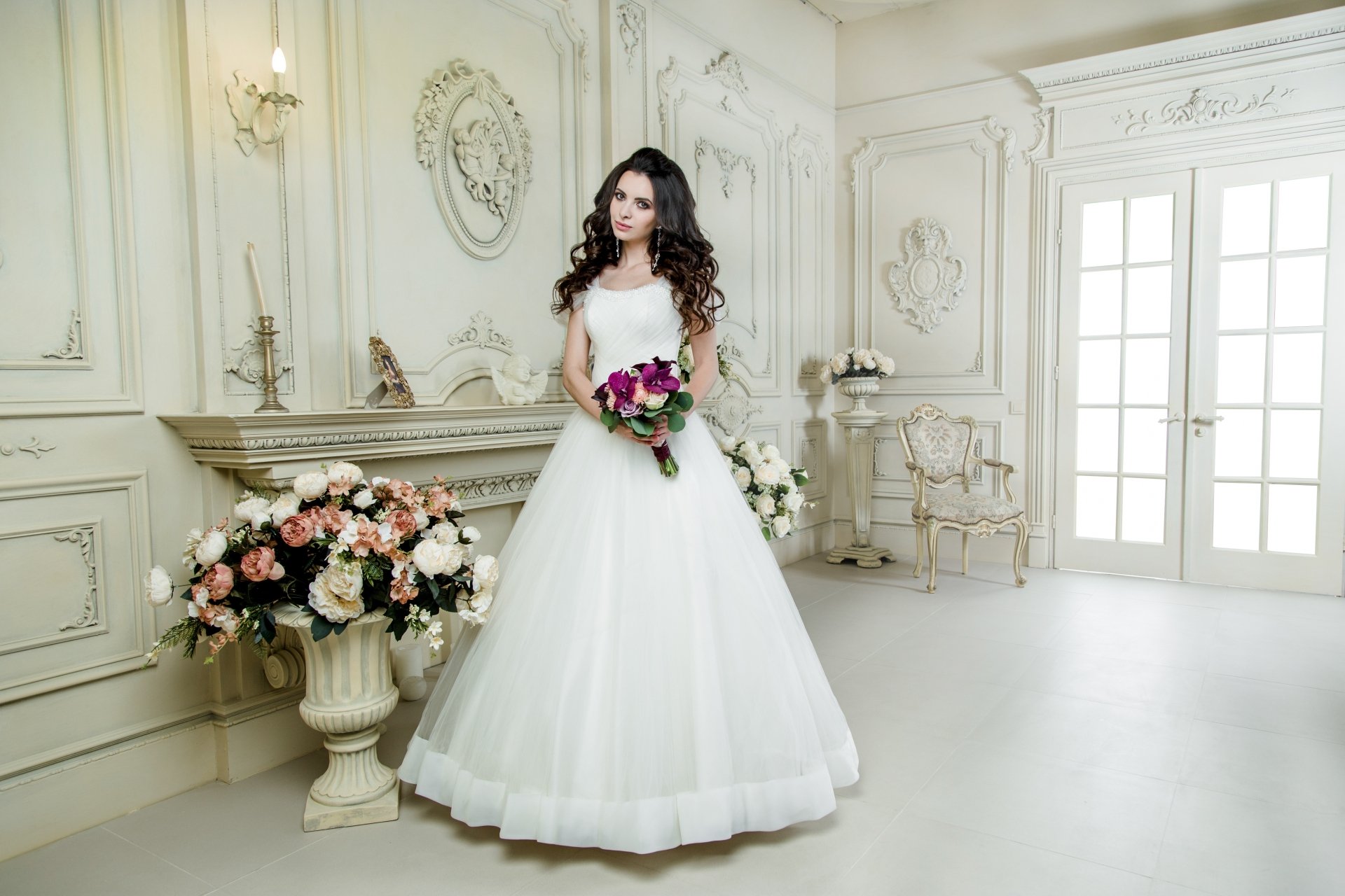 A brunette bride with brown eyes stands in an elegant room, wearing a white wedding dress and holding a vibrant bouquet, captured in stunning 4K Ultra HD detail.