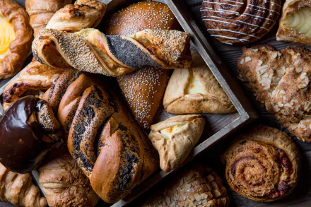 Close-up of assorted viennoiserie breakfast pastries—croissants, twists, rolls and danishes with glazes and seeds on a tray; baking textures rendered as a 2K Quad HD PC desktop wallpaper/background.