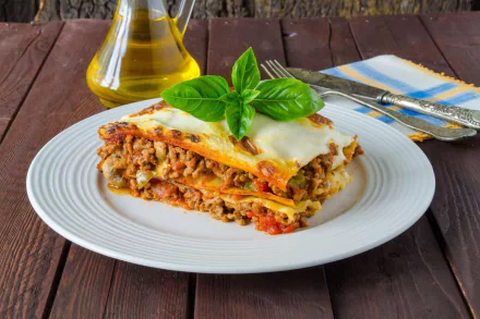 HD PC desktop wallpaper background: plated lasagna pasta food with melted cheese, meat sauce and basil garnish on a white plate, set on a wooden table with oil bottle and napkin.