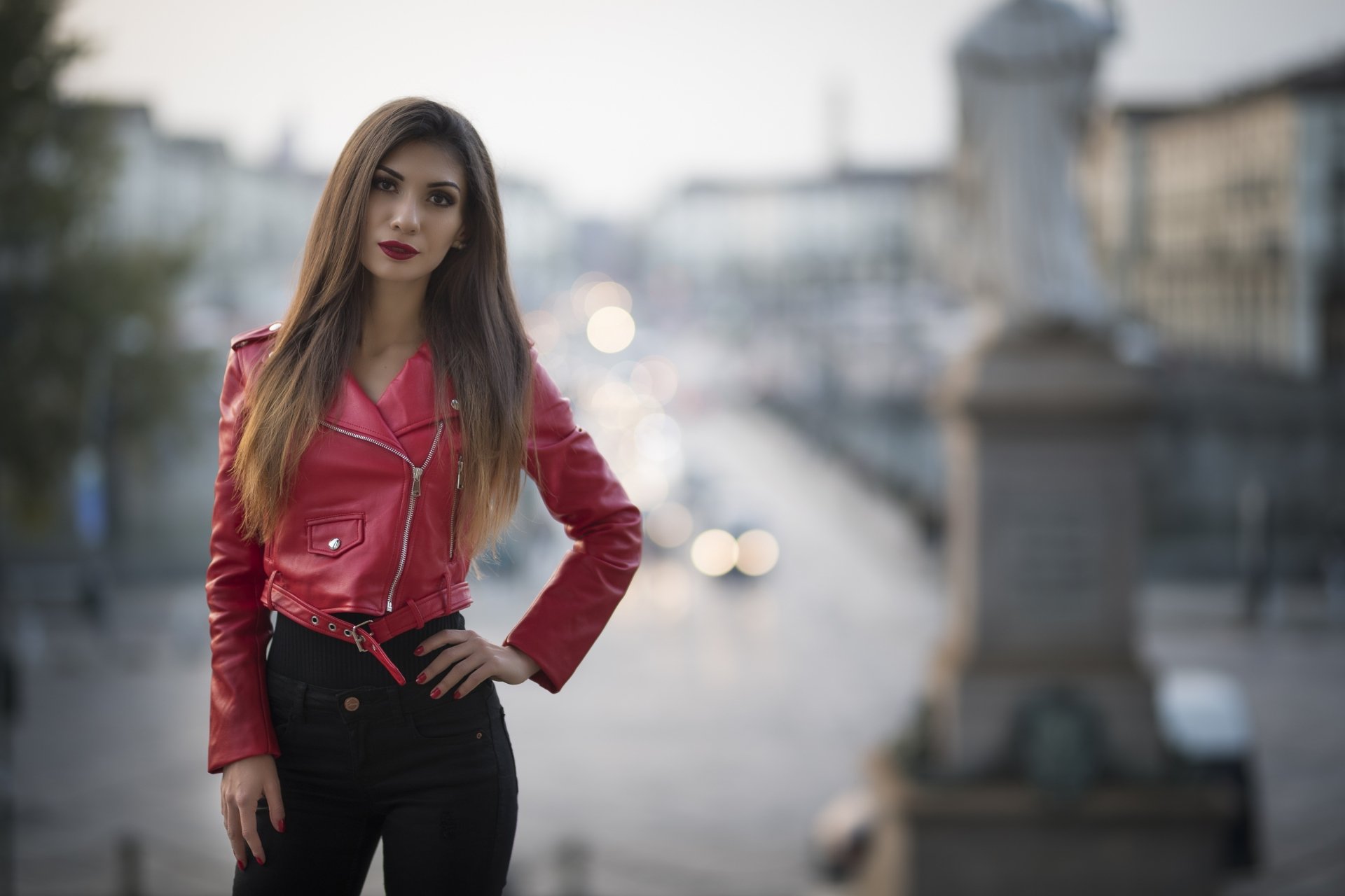 Brunette woman and model with long hair and brown eyes in a red leather jacket and lipstick, posed against a blurred city street (depth of field). 2K Quad HD PC desktop wallpaper.