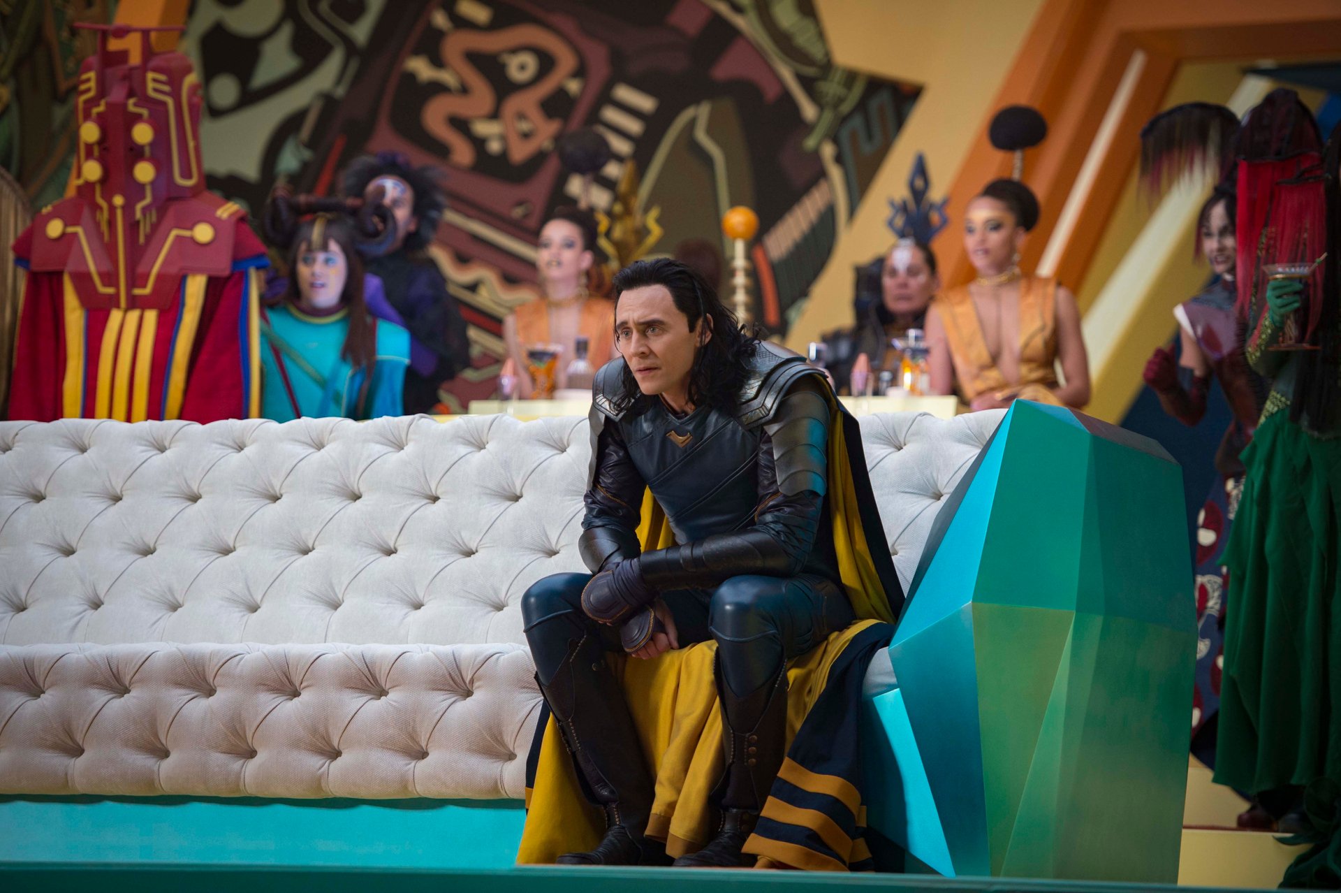 Loki (Tom Hiddleston) from Marvel's Thor: Ragnarok sits on a futuristic couch, surrounded by colorful characters, in this HD desktop wallpaper and background.