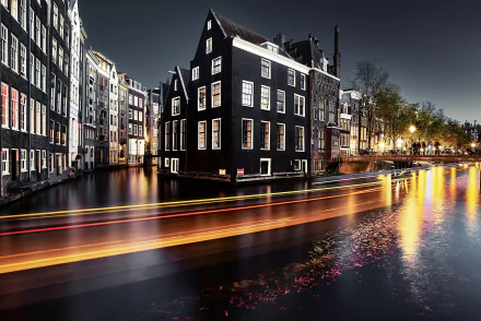 A stunning night view of Amsterdam's canals showcases iconic buildings and colorful boat trails, creating a dynamic time-lapse effect in this HD desktop wallpaper.