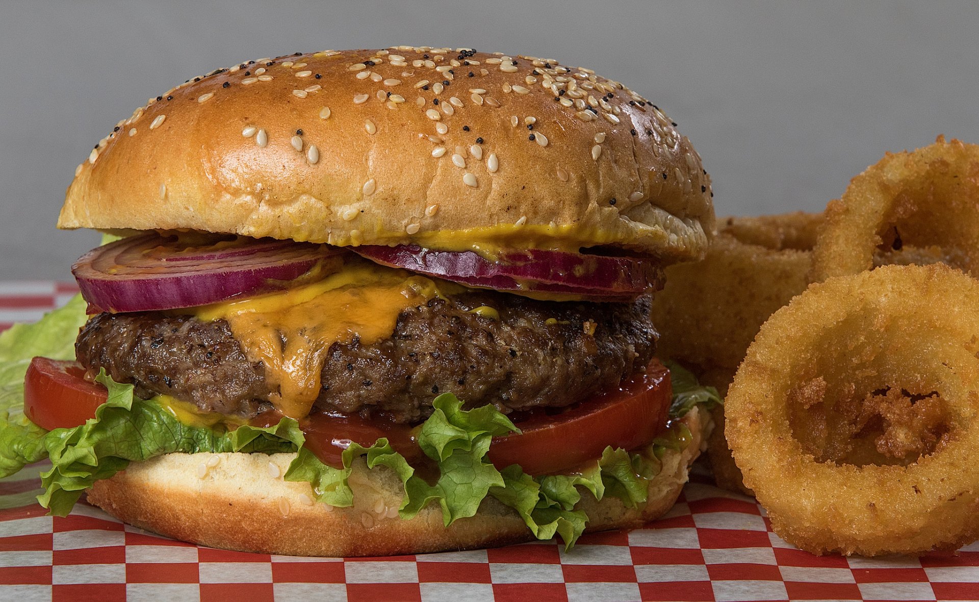 HD desktop wallpaper featuring a juicy cheeseburger with lettuce, tomato, and red onion, accompanied by crispy onion rings on a checkered paper background.