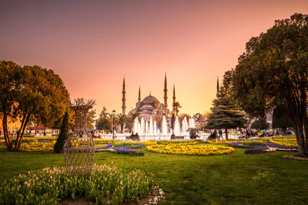 HD desktop wallpaper featuring the Blue Mosque (Sultan Ahmed Mosque) in Istanbul, Turkey, framed by a park with blooming flowers and a central fountain at sunset.