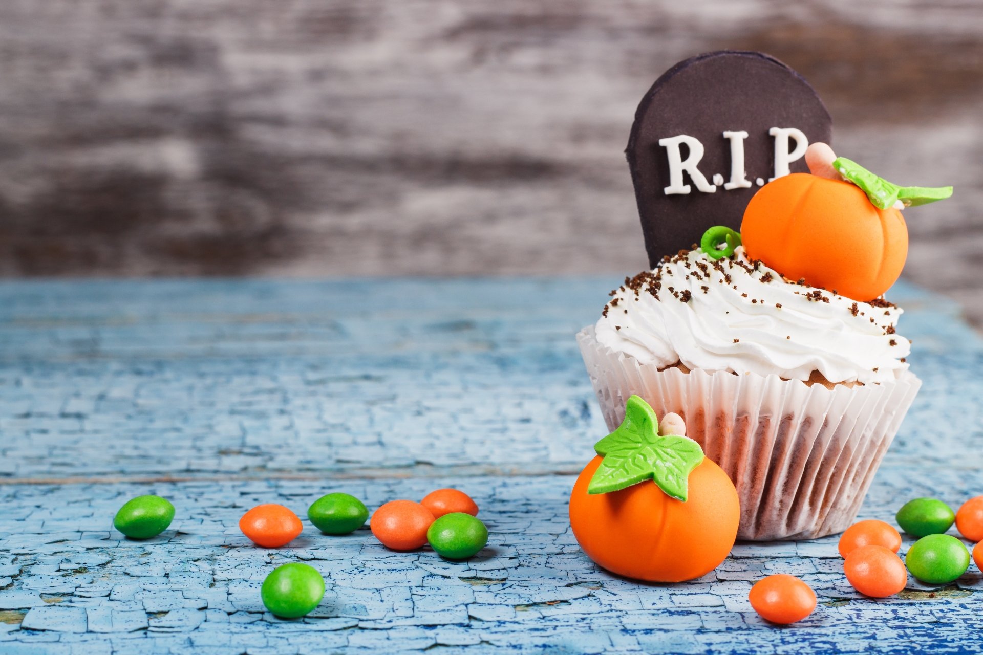 Halloween-themed cupcake with cream frosting, a chocolate R.I.P. tombstone, and small pumpkin candies on a cracked blue wooden surface, 4K Ultra HD wallpaper.