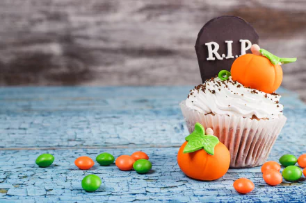 Halloween-themed cupcake with cream frosting, a chocolate R.I.P. tombstone, and small pumpkin candies on a cracked blue wooden surface, 4K Ultra HD wallpaper.