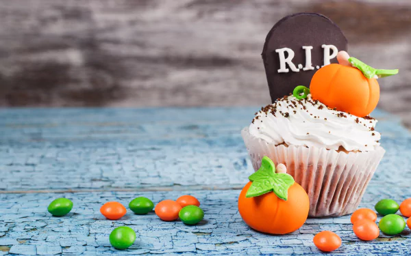 Halloween-themed cupcake with cream frosting, a chocolate R.I.P. tombstone, and small pumpkin candies on a cracked blue wooden surface, 4K Ultra HD wallpaper.