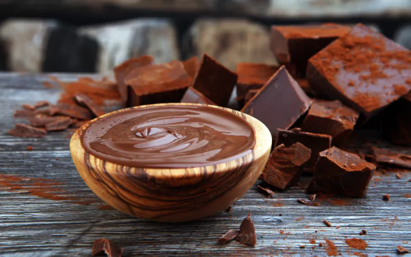 An 8K Ultra HD still life of creamy chocolate in a wooden bowl surrounded by chunks of chocolate and cocoa powder on a rustic wooden surface.
