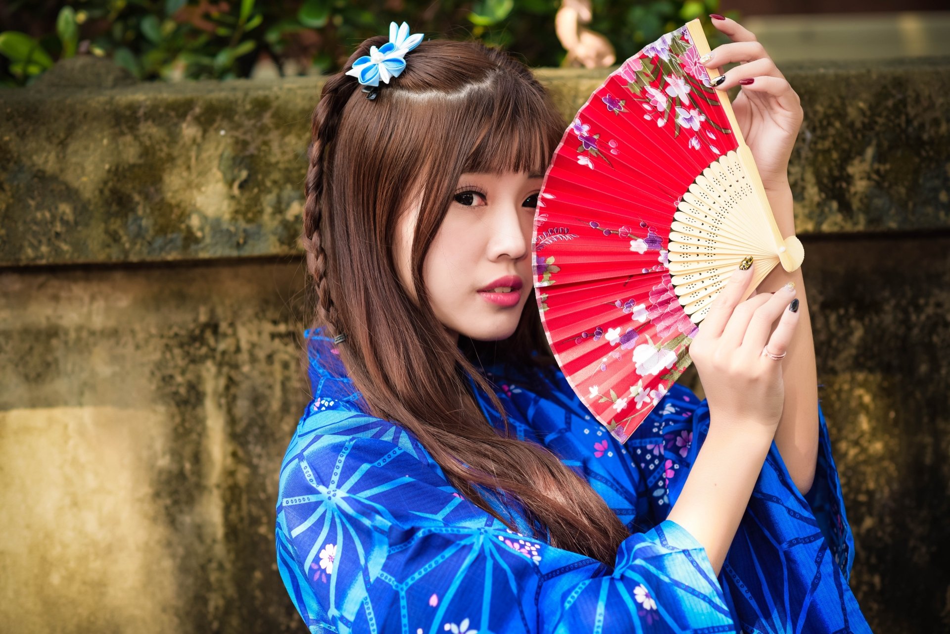 Asian woman with brown eyes and brunette braided hair wearing a blue kimono holds a red fan against a natural outdoor background.