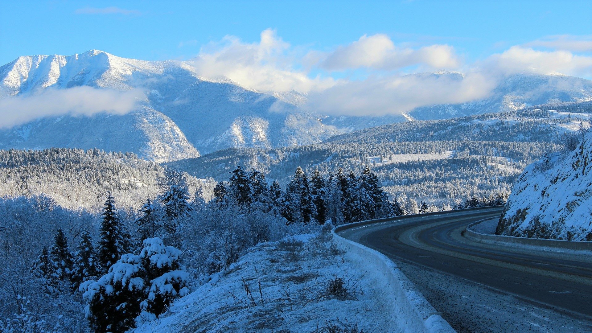 Snow-covered trees and a winding road lead through a winter landscape of snowy mountains under a bright blue sky, captured in HD for a desktop wallpaper.