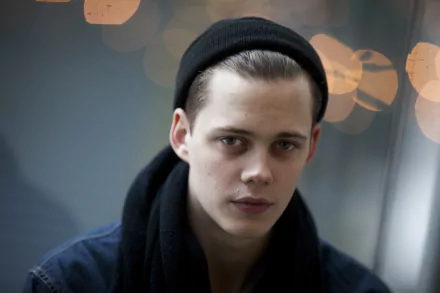 Close-up portrait of a young man in a dark beanie and scarf, serious expression against warm bokeh lights — moody 2K Quad HD PC desktop wallpaper.