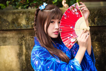 Asian woman with brown eyes and brunette braided hair wearing a blue kimono holds a red fan against a natural outdoor background.