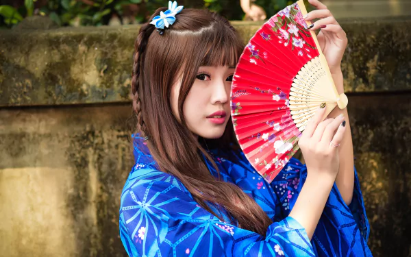Asian woman with brown eyes and brunette braided hair wearing a blue kimono holds a red fan against a natural outdoor background.