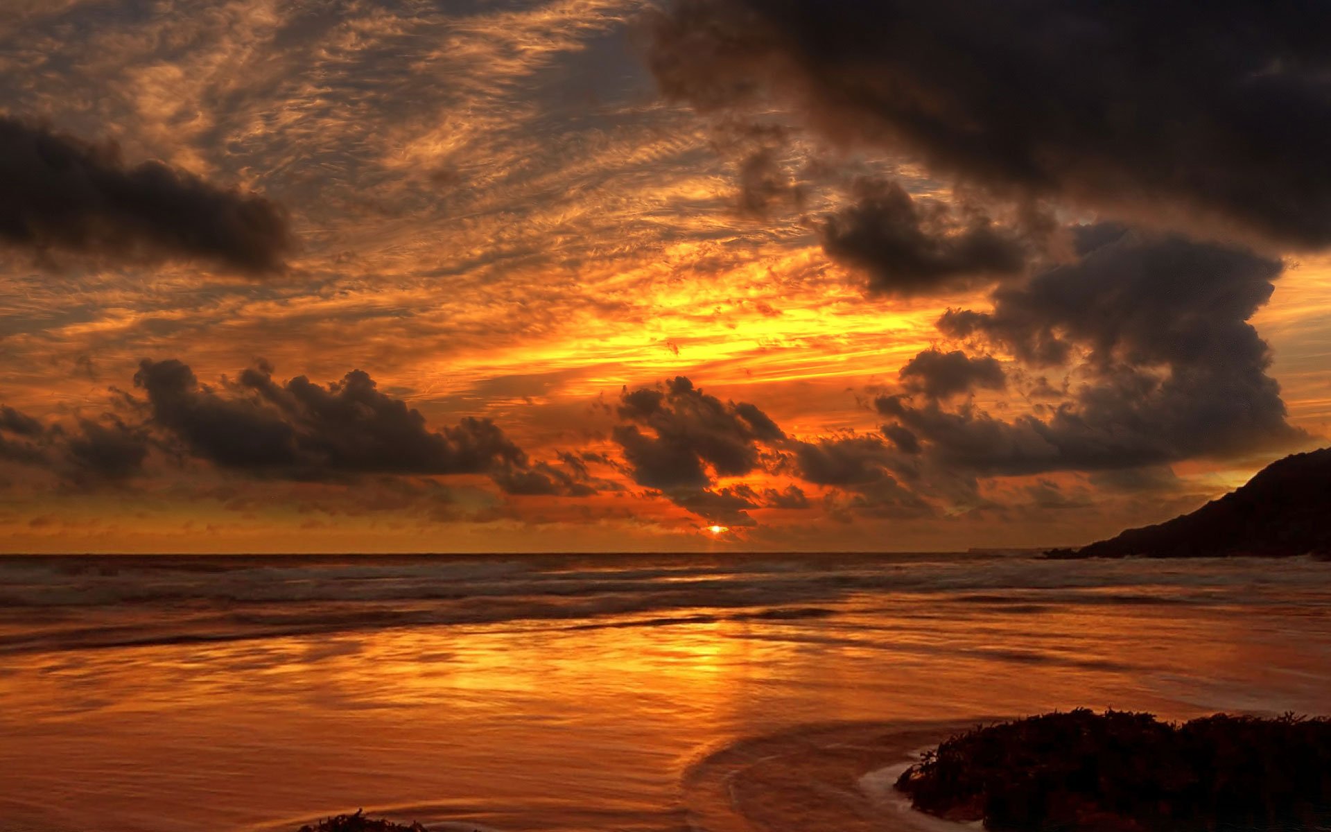 A stunning HD desktop wallpaper depicting a serene sunset over the ocean, with vibrant colors reflecting on the beach and dramatic clouds in the sky, creating a picturesque horizon.