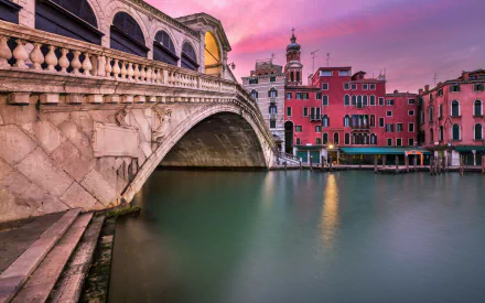 2K Quad HD desktop wallpaper: Venetian Rialto Bridge spanning a calm canal with colorful houses along the water at sunset, man-made stone bridge in Italy.