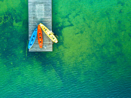 Aerial view of a wooden pier extending over clear green water with three colorful kayaks docked, captured in 4K Ultra HD resolution for a PC desktop wallpaper.