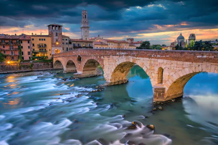 HD desktop wallpaper showcasing Ponte Pietra, a historic stone bridge over the river in Verona, Italy, with nearby architecture under a dramatic sky.
